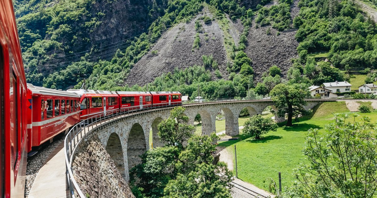 The red Bernina train crossing a stone viaduct between green meadows and wooded hills